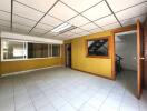 Spacious room with yellow walls, tiled floor, window, and wooden door, with adjacent room featuring staircase