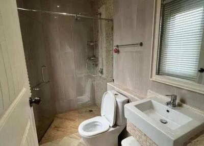 Modern bathroom with neutral tile walls, glass shower enclosure, toilet, sink, and a window with blinds