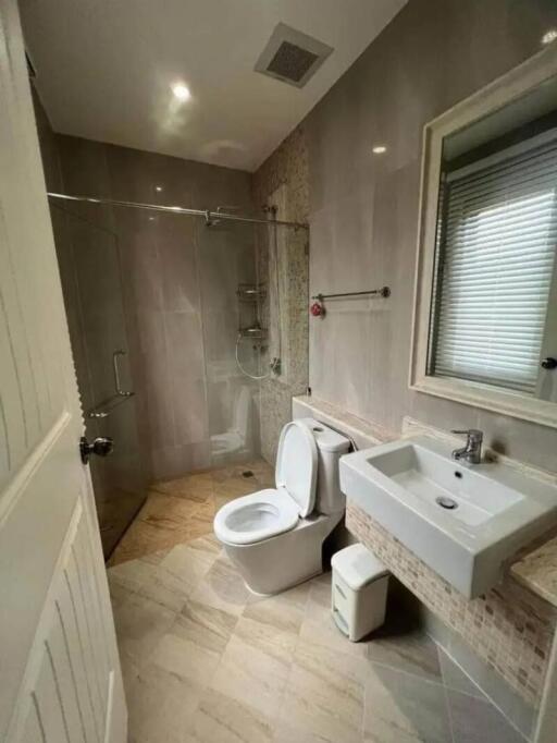 Modern bathroom with neutral tile walls, glass shower enclosure, toilet, sink, and a window with blinds