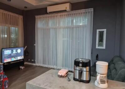 Well-lit living room with sheer curtains, air conditioner, TV, and kitchen appliances