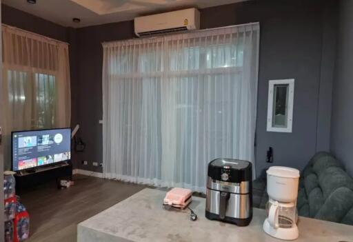 Well-lit living room with sheer curtains, air conditioner, TV, and kitchen appliances