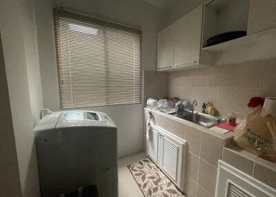 A compact laundry room featuring a washing machine, sink, storage cabinets, and a window with blinds