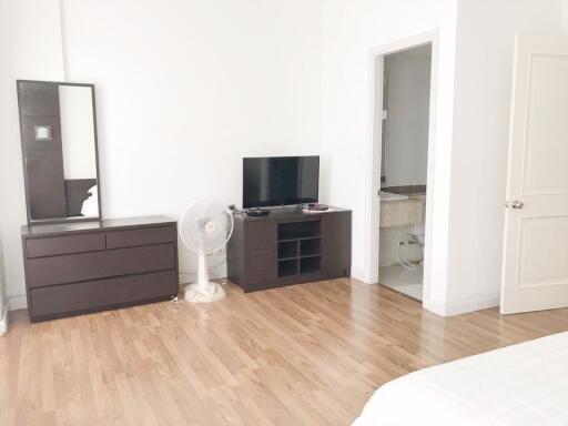 A bedroom with wooden flooring, dresser, mirror, fan, TV, and an attached bathroom