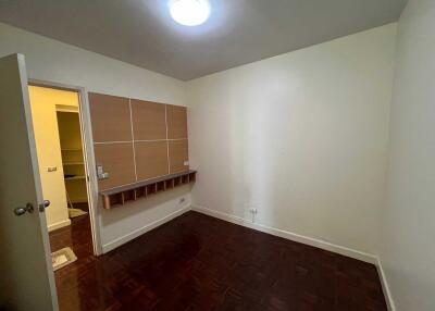 Empty bedroom with wooden floor, wall shelving, and a door to an illuminated adjacent space