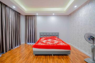 A well-lit bedroom with wooden flooring, a bed with a red and white bedspread, large curtains, and a ceiling fan.