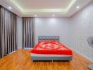 A well-lit bedroom with wooden flooring, a bed with a red and white bedspread, large curtains, and a ceiling fan.