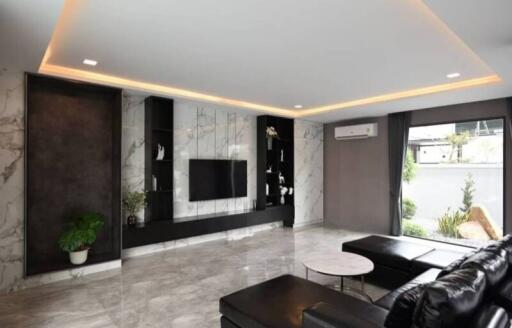 Modern living room with a marble accent wall, built-in black storage units, a flat-screen TV, a black sectional sofa, a small white coffee table, recessed lighting, and large windows with garden view.