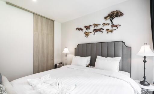 Modern bedroom with grey headboard, white bedding, two bedside lamps, and wall art featuring a tree and running horses