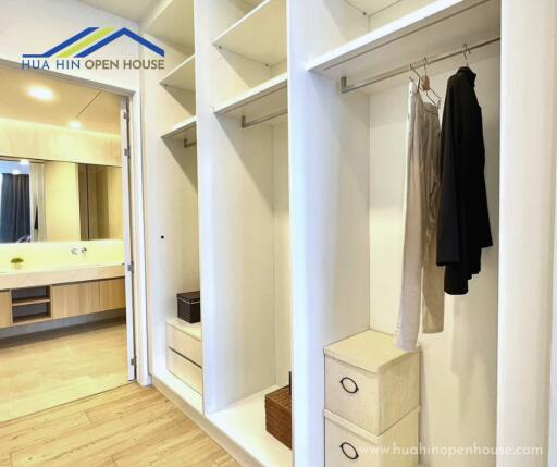 Walk-in closet with white shelves and hanging clothes, leading to a modern bathroom