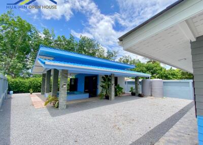 A single-story house with a blue roof and attached carport, surrounded by greenery and a gravel driveway