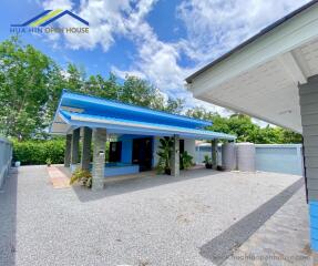 A single-story house with a blue roof and attached carport, surrounded by greenery and a gravel driveway