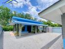 A single-story house with a blue roof and attached carport, surrounded by greenery and a gravel driveway