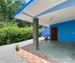 Exterior view of a property with a sheltered patio, blue exterior walls, greenery, and a gravel driveway