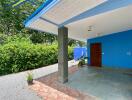 Exterior view of a property with a sheltered patio, blue exterior walls, greenery, and a gravel driveway