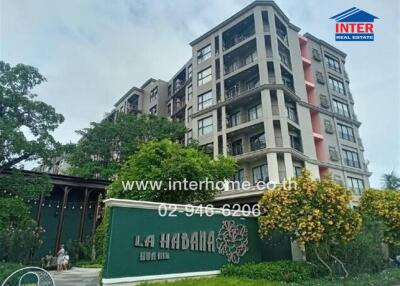 Exterior view of a multi-story residential building with greenery and a sign reading 