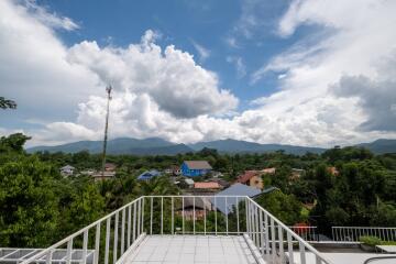 View from rooftop terrace overlooking trees, houses, and mountains under a partly cloudy sky