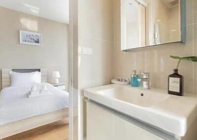 A view of a modern bathroom sink with a mirror and a bedroom with a neatly made bed in the background