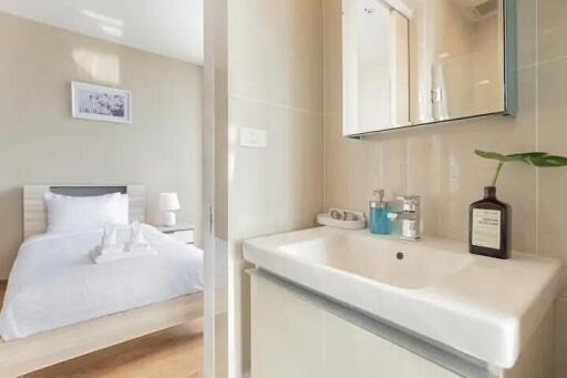 A view of a modern bathroom sink with a mirror and a bedroom with a neatly made bed in the background