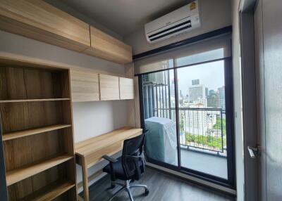 Small study area with a desk, chair, and shelving, adjacent to a balcony with a city view.