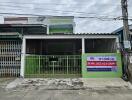 Front view of a residential building with a green gate and a 'for sale' sign