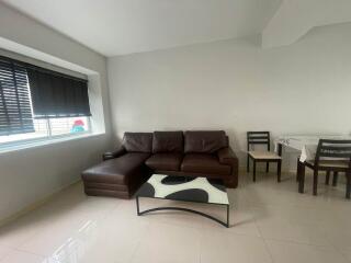 Living room with brown leather sectional sofa, glass coffee table, window with blinds, and adjacent dining area with table and chairs