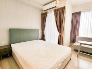 A cozy bedroom with an air conditioning unit, large bed, bedside tables, and natural light from large windows with curtains