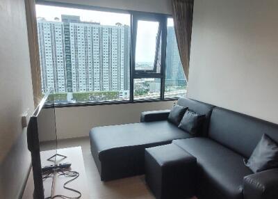 Modern living room with black leather sofa and large window overlooking high-rise buildings.