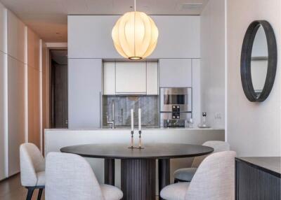 Modern dining area with round table, four chairs, elegant light fixture, and a view into a minimalist kitchen.