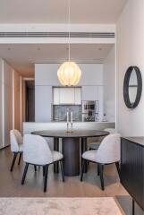 Modern dining area with round table, four chairs, elegant light fixture, and a view into a minimalist kitchen.