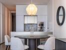 Modern dining area with round table, four chairs, elegant light fixture, and a view into a minimalist kitchen.