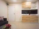 Compact kitchen area with wooden cabinets and dark countertops next to an entrance door, sofa, and round table with fruit