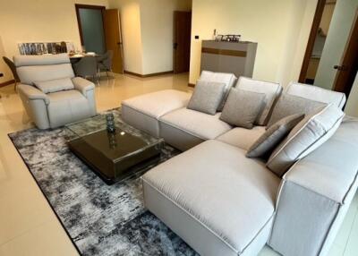 Modern living room with beige sofa and armchairs, glass coffee table, and dining area in the background