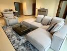 Modern living room with beige sofa and armchairs, glass coffee table, and dining area in the background