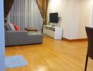 Living room with wooden flooring, gray sectional sofa, wall-mounted TV, white TV console, and large curtains