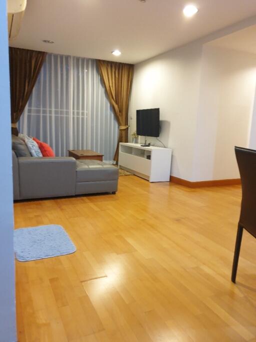 Living room with wooden flooring, gray sectional sofa, wall-mounted TV, white TV console, and large curtains