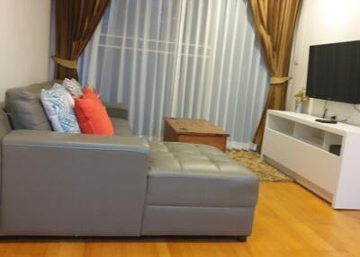 A modern living room with a gray sectional sofa, colorful pillows, wooden coffee table, TV on a white stand, and floor-to-ceiling curtains.