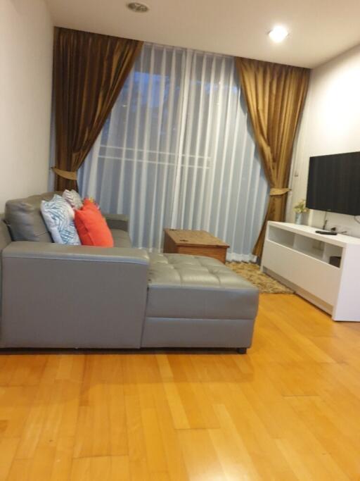 A modern living room with a gray sectional sofa, colorful pillows, wooden coffee table, TV on a white stand, and floor-to-ceiling curtains.