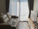 Living room with beige leather sofa, large window with sheer curtains, and wall-mounted TV