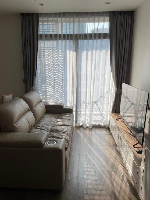 Living room with beige leather sofa, large window with sheer curtains, and wall-mounted TV