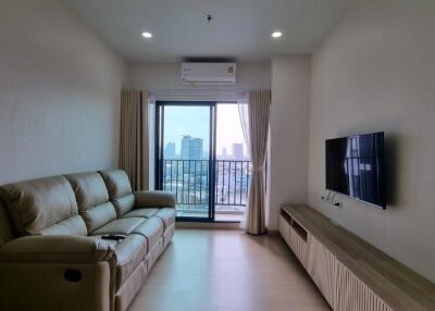 A cozy living room with a beige sofa, mounted TV, and large windows leading to a balcony with a city view