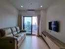 A cozy living room with a beige sofa, mounted TV, and large windows leading to a balcony with a city view