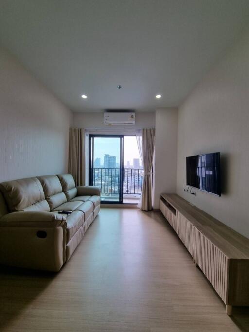 A cozy living room with a beige sofa, mounted TV, and large windows leading to a balcony with a city view