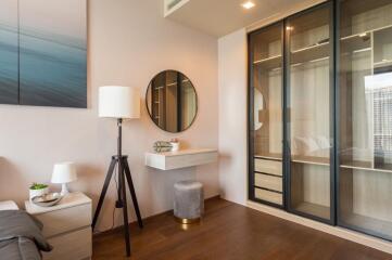 Modern bedroom with a minimalist design featuring a lamp, a round mirror, a small floating desk, a bedside table, a stool, and a built-in wardrobe with glass sliding doors.