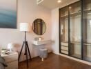 Modern bedroom with a minimalist design featuring a lamp, a round mirror, a small floating desk, a bedside table, a stool, and a built-in wardrobe with glass sliding doors.