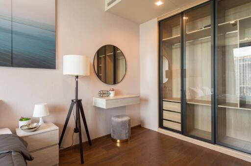 Modern bedroom with a minimalist design featuring a lamp, a round mirror, a small floating desk, a bedside table, a stool, and a built-in wardrobe with glass sliding doors.