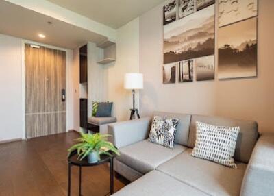 Modern living room with a light gray sectional sofa, decorative pillows, wall art, a floor lamp, and a small round table with a plant