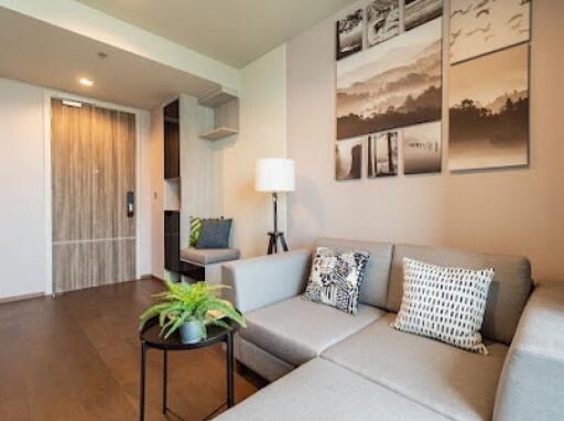 Modern living room with a light gray sectional sofa, decorative pillows, wall art, a floor lamp, and a small round table with a plant