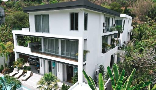 Modern white multi-story house with large windows and balconies, surrounded by lush vegetation