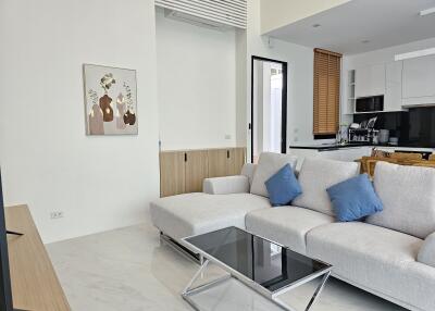 Modern living room with a light gray sectional sofa and blue cushions, large wall artwork, glass-top coffee table, and a view into the kitchen area.