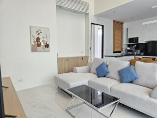 Modern living room with a light gray sectional sofa and blue cushions, large wall artwork, glass-top coffee table, and a view into the kitchen area.